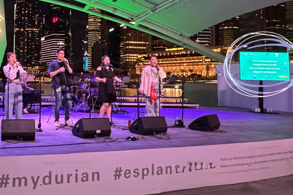 The photo captures a live musical performance at an outdoor venue, likely the Esplanade in Singapore, with the city skyline illuminated in the background. Four performers, three women and one man, stand on stage, each holding a microphone and singing. A live band setup is visible in the background with drums and a keyboard.

To the right of the stage, a large digital screen displays song lyrics: “You can dance, you can jive, having the time of your life. Ooh, see that girl, watch that scene, digging the dancing queen.” This screen ensures accessibility, enabling the audience to follow the lyrics visually. The foreground shows part of the stage branding with the text “#my.durian #esplanade.”

This image reflects an effort towards inclusivity by incorporating visible song lyrics, enhancing the experience for all attendees, including deaf and hard of hearing.